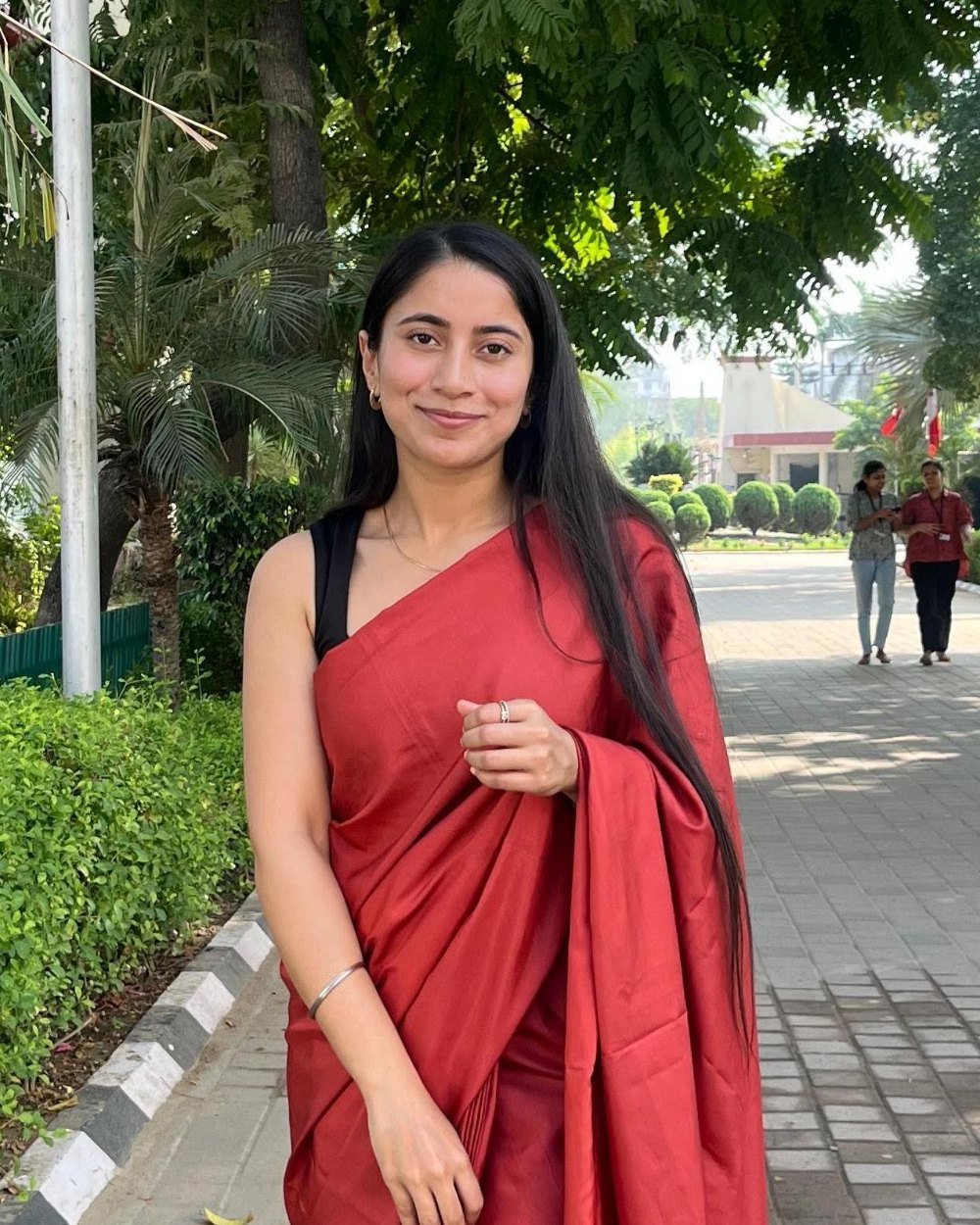 Gurleen Kaur, smiling, in a red sari on a tree-lined campus path.